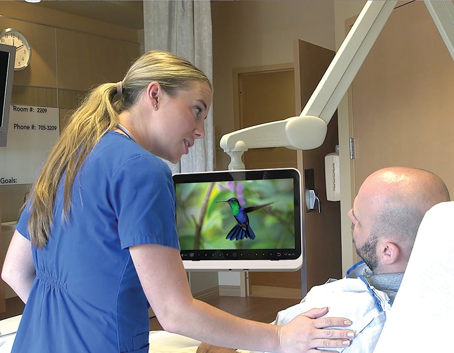 A nurse helping a patient in a hospital bed, showing educational videos on a medTV16 by PDi, mounted to the wall with a PDi arm.