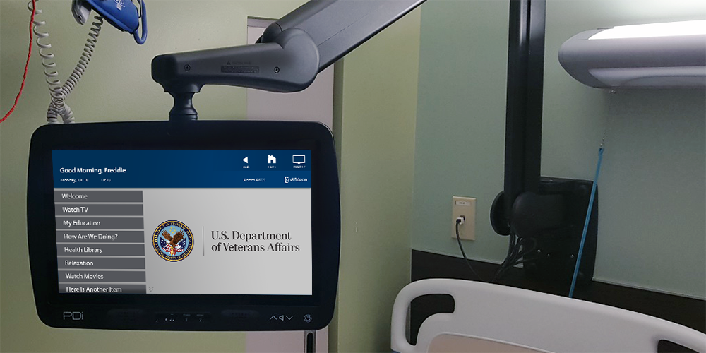 A medTV19 by PDi next to a patient bed at a Veterans Hospital displaying a welcome screen, mounted to the wall with a PDi arm.