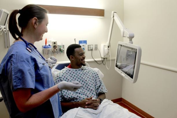 A nurse holding a remote and showing a patient in a hospital bed the content on a medTV19 by PDi, mounted to the wall with a PDi arm.